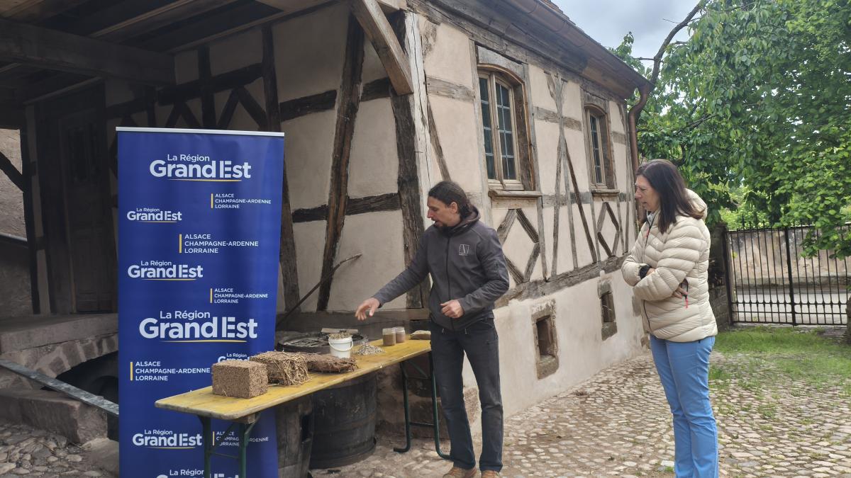 Deux personnes discutent devant une maison traditionnelle à colombages aux murs en torchis, typique du massif des Vosges. À gauche, Jacques Bruderer se tient près d’une table en bois sur laquelle sont disposés des échantillons de matériaux biosourcés. À droite,Denise Buhl, conseillère régionale, l’écoute attentivement.